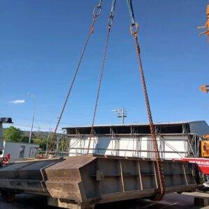 Ein großer Metallcontainer wird von einem Kran angehoben und stellt die beeindruckende Ingenieurskunst von Stahlwasserbau unter Beweis. Der Container hängt frei vor einem klaren blauen Himmel, im Hintergrund sind Industriegebäude und Stromleitungen zu sehen.