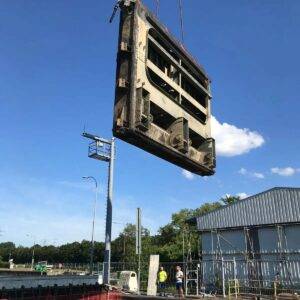 Inmitten eines klaren blauen Himmels und üppigen Grüns wird eine große Metallkonstruktion, Teil eines komplexen Schleusenbau-Projekts, von einem Kran auf die Baustelle gehoben. Zwei Arbeiter mit Helmen beobachten das Ganze von unten, während sie neben einem teilweise fertiggestellten Gebäude stehen.