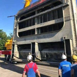 Arbeiter in Sicherheitsausrüstung beobachten, wie eine große, rechteckige Metallkonstruktion, die für Stahlwasserbau-Projekte von entscheidender Bedeutung ist, von einem Kran auf einem Tieflader transportiert wird. Im Hintergrund ist vor einem klaren blauen Himmel ein gelber Kran teilweise zu erkennen, der fortschrittliche Metallverarbeitungstechniken hervorhebt.