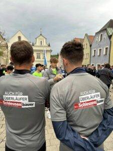 Zwei Personen in grauen Hemden mit dem Text „AUCH FÜR ÜBERLÄUFER Die Schleusenbauer“ stehen mit dem Rücken zur Kamera und demonstrieren ihr Fachwissen in der CNC-Fertigung. Eine Gruppe von Menschen hat sich auf einem Stadtplatz versammelt, im Hintergrund sind farbenfrohe Gebäude zu sehen, die eine Szenerie voller lokaler Kultur und Handwerkskunst hervorheben.