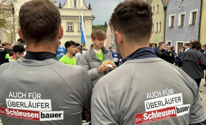 Zwei Personen in grauen Hemden mit dem Text „AUCH FÜR ÜBERLÄUFER Die Schleusenbauer“ stehen mit dem Rücken zur Kamera und demonstrieren ihr Fachwissen in der CNC-Fertigung. Eine Gruppe von Menschen hat sich auf einem Stadtplatz versammelt, im Hintergrund sind farbenfrohe Gebäude zu sehen, die eine Szenerie voller lokaler Kultur und Handwerkskunst hervorheben.