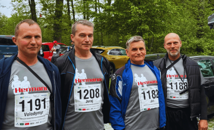 Vier Männer in passenden grauen T-Shirts mit Startnummern posieren gemeinsam im Freien auf einem gepflasterten Weg und heben die Teamarbeit hervor, die der Präzision in der Metallverarbeitung ähnelt. Sie sind von Grünflächen und geparkten Autos im Hintergrund umgeben, was an einen Marathon oder eine Laufveranstaltung erinnert.