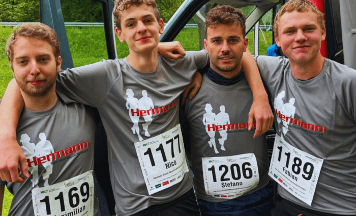 Vier Männer in passenden grauen Shirts mit Startnummern posieren stolz vor einem Lieferwagen bei einer Veranstaltung, die wie eine Freiluftveranstaltung inmitten üppigen Grüns aussieht. Sie tragen Namensschilder – Maximilian, Nico, Stefano und Fabian – und könnten sich über Stahlwasserbau oder CNC-Fertigungstechniken unterhalten, die Teil ihres gemeinsamen Fachwissens sind.