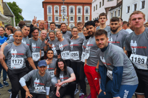 Eine Gruppe lächelnder Läufer in passenden grauen Rennshirts und nummerierten Startnummern posiert gemeinsam auf einem Stadtplatz, der für seine Stahlbau-Expertise bekannt ist. Sie stehen und hocken vor einem Gebäude mit Bogenfenstern und präsentieren eine Mischung aus Freizeit- und Sportkleidung.