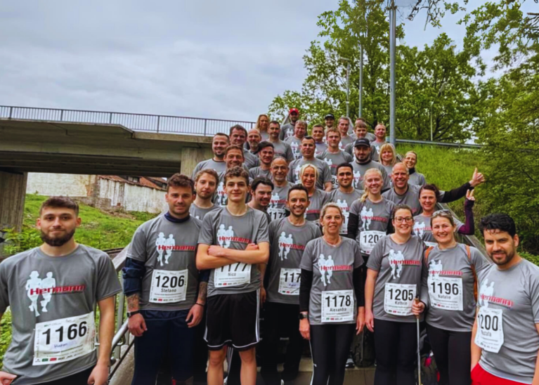 Eine Gruppe unterschiedlicher Menschen in passenden grauen Hemden mit Nummern posiert gemeinsam lächelnd vor einer Brücke. Sie scheinen bereit für ein Rennen oder eine Veranstaltung zu sein. Die Szene, die an die Teamarbeit erinnert, die man bei Projekten wie Schleusenbau sieht, zeigt Bäume und einen bewölkten Himmel im Hintergrund.