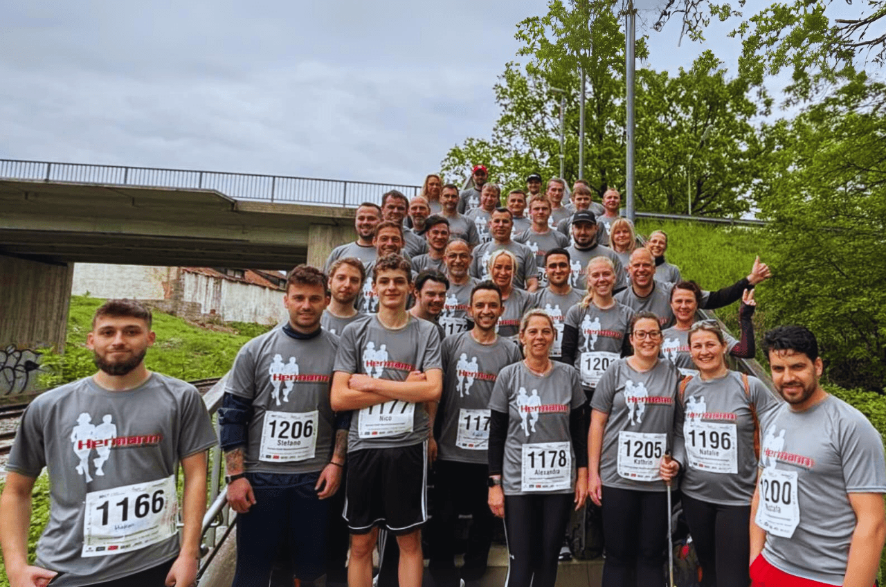 Eine Gruppe unterschiedlicher Menschen in passenden grauen Hemden mit Nummern posiert gemeinsam lächelnd vor einer Brücke. Sie scheinen bereit für ein Rennen oder eine Veranstaltung zu sein. Die Szene, die an die Teamarbeit erinnert, die man bei Projekten wie Schleusenbau sieht, zeigt Bäume und einen bewölkten Himmel im Hintergrund.