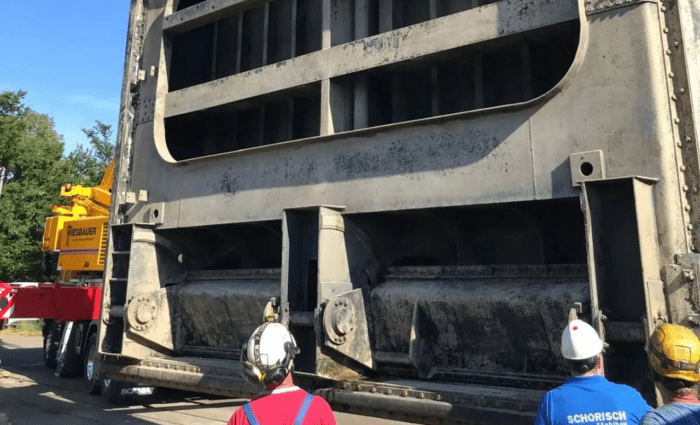 Arbeiter mit Helmen beobachten, wie eine große, industrielle Metallkonstruktion von einem gelben Kran transportiert wird. Die Szene spielt im Freien, und auf dem Hemd eines Arbeiters sind die Worte „SCHORISCH“ und „Stahlbau“ zu sehen. Diese Demonstration der Präzision im Stahlwasserbau findet vor einer üppigen Kulisse aus Bäumen statt.