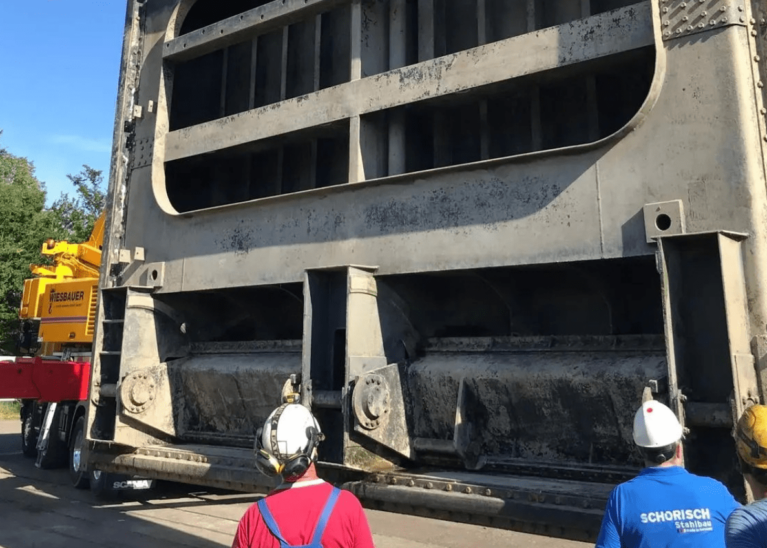 Arbeiter mit Helmen beobachten, wie eine große, industrielle Metallkonstruktion von einem gelben Kran transportiert wird. Die Szene spielt im Freien, und auf dem Hemd eines Arbeiters sind die Worte „SCHORISCH“ und „Stahlbau“ zu sehen. Diese Demonstration der Präzision im Stahlwasserbau findet vor einer üppigen Kulisse aus Bäumen statt.