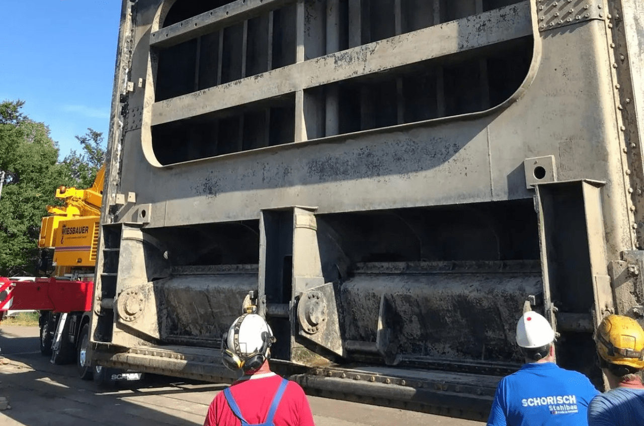 Arbeiter mit Helmen beobachten, wie eine große, industrielle Metallkonstruktion von einem gelben Kran transportiert wird. Die Szene spielt im Freien, und auf dem Hemd eines Arbeiters sind die Worte „SCHORISCH“ und „Stahlbau“ zu sehen. Diese Demonstration der Präzision im Stahlwasserbau findet vor einer üppigen Kulisse aus Bäumen statt.