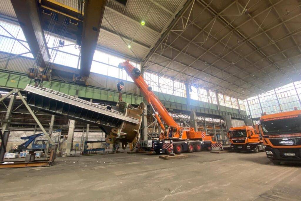 Ein großer orangefarbener Kran in einem Industrielager hebt schweres Gerät. In der Nähe stehen zwei orangefarbene Lastwagen. Das Gebäude ist geräumig, hat eine gewölbte Decke und große Fenster, die für natürliches Licht sorgen.