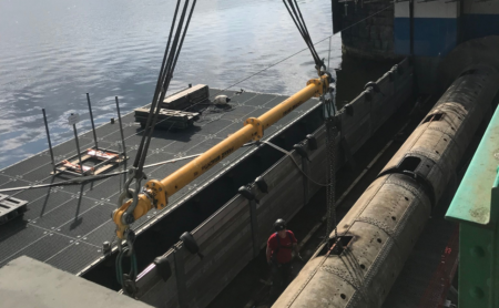 Auf einer Baustelle über dem Wasser sind schwere Maschinen im Einsatz. Ein großer gelber Balken, der für das Schleusenbau-Projekt von entscheidender Bedeutung ist, wird von einem Kran herabgelassen, während ein Arbeiter mit Schutzhelm daneben steht. Im Hintergrund sind Gerüste und Geräte zu sehen.