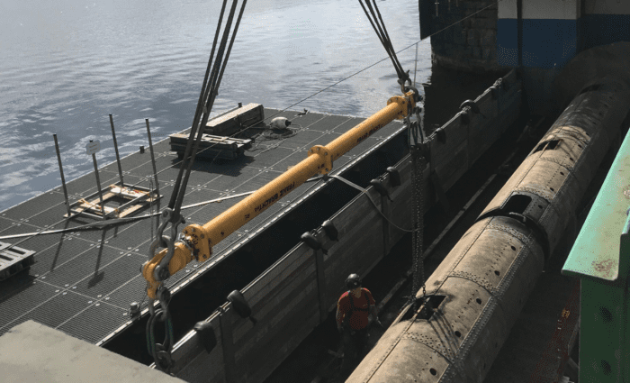 Auf einer Baustelle über dem Wasser sind schwere Maschinen im Einsatz. Ein großer gelber Balken, der für das Schleusenbau-Projekt von entscheidender Bedeutung ist, wird von einem Kran herabgelassen, während ein Arbeiter mit Schutzhelm daneben steht. Im Hintergrund sind Gerüste und Geräte zu sehen.