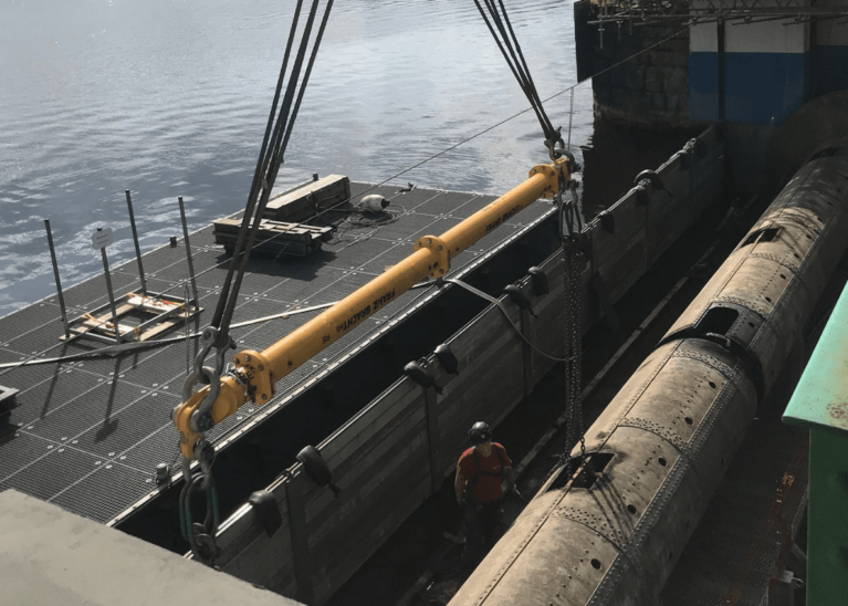 Auf einer Baustelle über dem Wasser sind schwere Maschinen im Einsatz. Ein großer gelber Balken, der für das Schleusenbau-Projekt von entscheidender Bedeutung ist, wird von einem Kran herabgelassen, während ein Arbeiter mit Schutzhelm daneben steht. Im Hintergrund sind Gerüste und Geräte zu sehen.