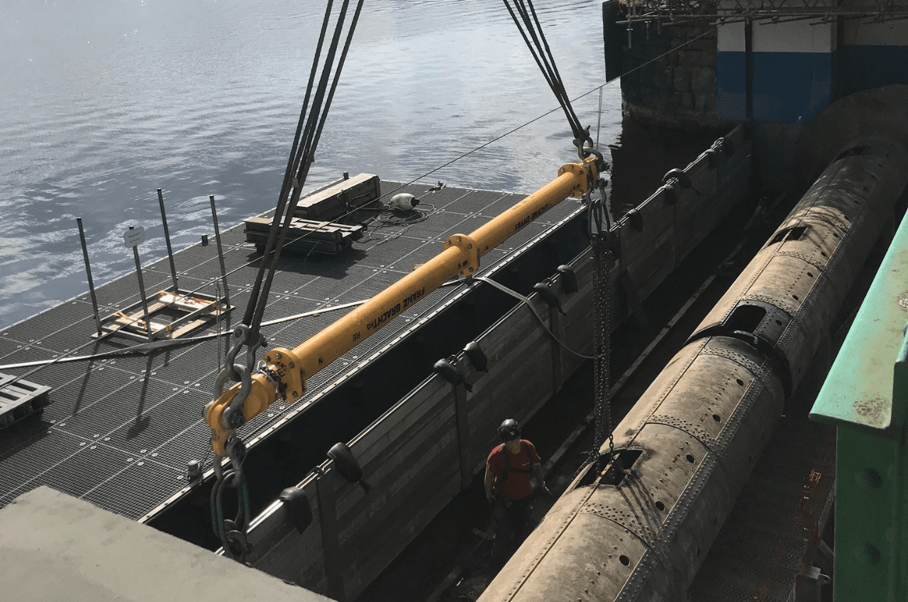 Auf einer Baustelle über dem Wasser sind schwere Maschinen im Einsatz. Ein großer gelber Balken, der für das Schleusenbau-Projekt von entscheidender Bedeutung ist, wird von einem Kran herabgelassen, während ein Arbeiter mit Schutzhelm daneben steht. Im Hintergrund sind Gerüste und Geräte zu sehen.