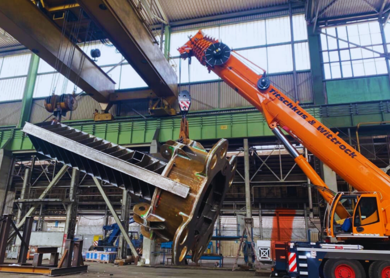 In der Fabrik arbeiten ein leuchtend orangefarbener Kran und ein Deckenkran in perfekter Harmonie zusammen, um ein schweres Industriebauteil anzuheben, und zeigen damit die Präzision, die für Hermann Maschinenbau charakteristisch ist. Die industrielle Umgebung mit hohen Decken lässt auf einen umfangreichen Bau- oder Fertigungsbetrieb schließen.