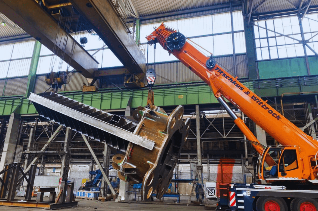 In der Fabrik arbeiten ein leuchtend orangefarbener Kran und ein Deckenkran in perfekter Harmonie zusammen, um ein schweres Industriebauteil anzuheben, und zeigen damit die Präzision, die für Hermann Maschinenbau charakteristisch ist. Die industrielle Umgebung mit hohen Decken lässt auf einen umfangreichen Bau- oder Fertigungsbetrieb schließen.